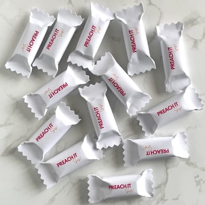 Puede incluir: Una colección de envoltorios de caramelos blancos con el texto "PREACH IT Sis!" impreso en rosa en cada envoltorio. Los envoltorios están esparcidos sobre una superficie de mármol blanco.