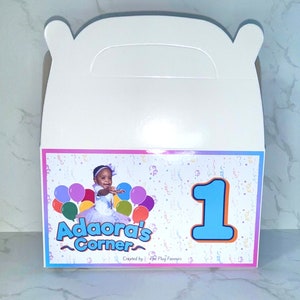 May include: A white favor box with a pink, white, and blue design. The box has a handle and features a photo of a young girl in a blue dress with a crown. The box also has the text "Adaora's Corner" and the number "1" in a blue circle.