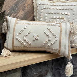 May include: Two decorative throw pillows with a beige linen fabric and white shell accents. The pillows have fringe trim and tassels.