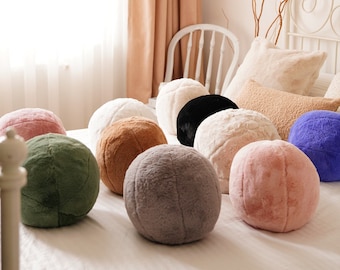 Puede incluir: Una colección de nueve almohadas redondas y mullidas en varios colores, incluyendo rosa, verde, marrón, gris, blanco y azul. Las almohadas están dispuestas sobre una colcha blanca.