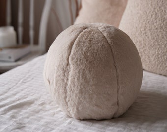 Puede incluir: Una almohada redonda y esponjosa de color crema con una costura en el centro, sentada sobre una colcha blanca.