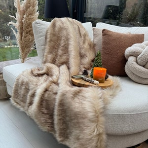 Può includere: Una coperta in pelliccia sintetica marrone drappeggiata su un divano bianco con un cuscino marrone e un cuscino bianco con un design a nodo. Un piccolo vassoio di legno con una candela e una piccola decorazione di pino si trova sul divano.