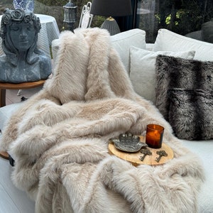 May include: A beige faux fur throw blanket draped over a white sofa with a black and white patterned cushion. A wooden tray with a silver hand-shaped dish, a candle, and two keys is on the blanket.