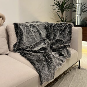 Op de afbeelding: Een luxe imitatiebont deken in grijs en zwart is over de arm van een beige bank gedrapeerd. De textuur van de deken is prominent aanwezig, wat een gezellige uitstraling creëert. De deken voegt een vleugje luxe toe aan de woonkamer.