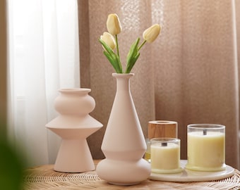 Puede incluir: Un jarrón de cerámica rosa claro con una base ancha y un cuello estrecho contiene un ramo de tulipanes amarillo pálido. Dos velas en portavelas de vidrio transparente y un portavelas de madera están sobre una mesa de mimbre tejido.