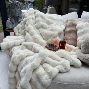 Puede incluir: Una manta de piel sintética de color marfil, sobre un sofá curvo. La manta tiene una apariencia texturizada y ondulada. Se ven cojines decorativos en tonos crema y beige. Una pequeña vela y una bandeja decorativa están en el sofá.