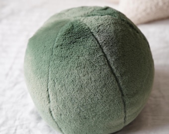 May include: A round, plush, sage green decorative pillow. The pillow has a soft, textured surface and is divided into sections by stitched seams. The background is a white, wrinkled fabric.
