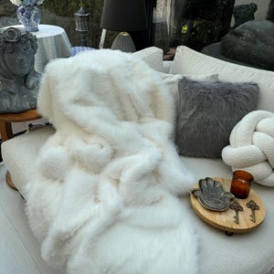 Può includere: Una coperta in pelliccia sintetica bianca drappeggiata su un divano bianco con un cuscino in pelliccia sintetica grigia e un cuscino bianco annodato. Un vassoio decorativo con un piatto argentato a forma di mano e chiavi si trova sul divano.