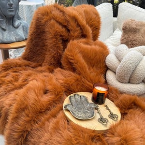 May include: A plush, rust-colored faux fur throw blanket draped over a sofa. A wooden tray holds a decorative hand-shaped dish, two antique keys, and a lit candle. Decorative pillows and a statue are in the background.