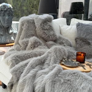 Puede incluir: Una manta de piel sintética gris sobre un sofá blanco. La manta de felpa es el punto focal, con una apariencia suave y texturizada. Cojines decorativos y una bandeja de madera con una vela y llaves completan la acogedora escena de la sala de estar.