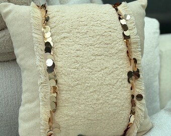 May include: Cream-colored decorative pillow with a textured center and fringed edges. Two vertical rows of gold sequins adorn the pillow, adding a touch of glamour. The pillow is square-shaped and appears to be made of soft fabric.
