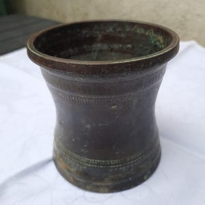 May include: A dark bronze mortar with a flared rim and base, featuring a cylindrical body with a slightly concave centre. The exterior surface displays a weathered patina with visible green oxidation and two horizontal bands of decorative etching.