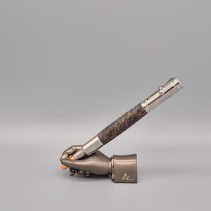 Puede incluir: Un bolígrafo de lujo con tapa y detalles de metal plateado, con un cuerpo con un patrón de vetas de madera multicolor único. El bolígrafo descansa sobre un portalápices metálico con forma de mano. El portalápices tiene la letra "A".