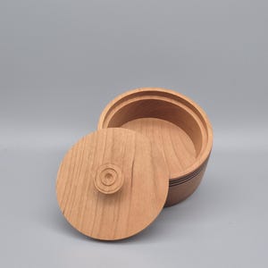 May include: A light-coloured wooden box with a lid, set against a grey background. The lid is slightly ajar, revealing the interior of the box. The box has a circular shape with a decorative design.