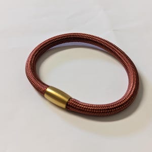 Op de afbeelding: Een bordeauxrode haarband met een geweven textuur. Het haaraccessoire heeft een goudkleurige metalen sluiting. De haarband is cirkelvormig en ligt op een wit oppervlak.