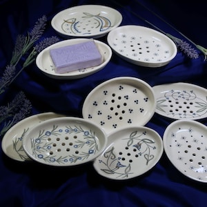 Può includere: Una collezione di portasapone ovali in ceramica con decorazioni floreali e geometriche blu e verdi. Un portasapone contiene una saponetta alla lavanda. I portasapone sono disposti su un tessuto blu scuro.