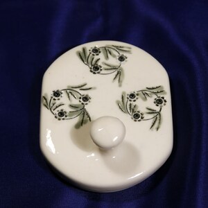 May include: White ceramic lid with a knob and three green floral designs with black centers.