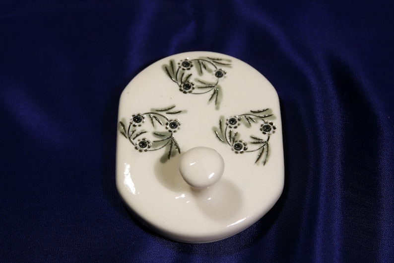 May include: White ceramic lid with a knob and three green floral designs with black centers.