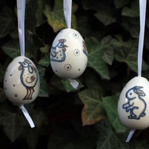 Puede incluir: Tres adornos de huevo de Pascua de cerámica blanca con diseños pintados en azul y marrón de un gallo, una gallina y un conejo. Cada huevo cuelga de una cinta blanca sobre un fondo de hojas verdes.