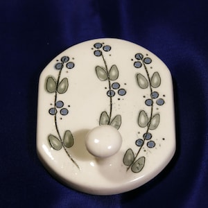 May include: White ceramic hook with a floral design in shades of blue and green. The hook is shaped like a rounded rectangle and has a single hook in the center.