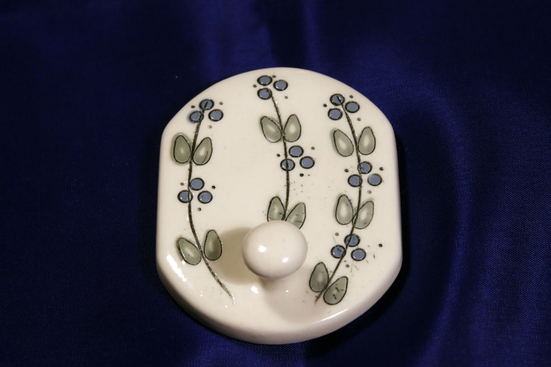 May include: White ceramic hook with a floral design in shades of blue and green. The hook is shaped like a rounded rectangle and has a single hook in the center.