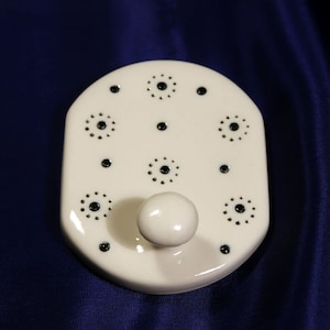 May include: White ceramic hook with a single hook and a decorative pattern of black circles. The hook is designed to hang items on a wall.