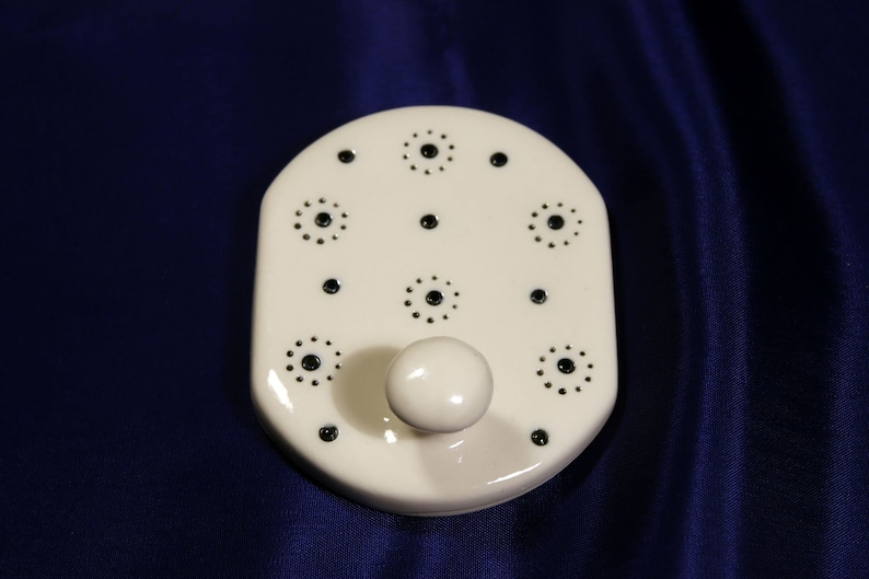 May include: White ceramic hook with a single hook and a decorative pattern of black circles. The hook is designed to hang items on a wall.