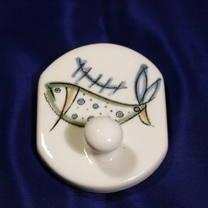 May include: White ceramic hook with a stylized fish design in blue, green, and brown. The fish has a dotted pattern and is swimming over a fence-like design.