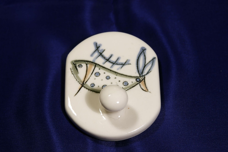 May include: White ceramic hook with a stylized fish design in blue, green, and brown. The fish has a dotted pattern and is swimming over a fence-like design.