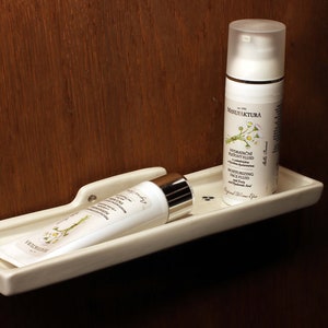 May include: A white ceramic shelf displays two skincare products against a dark wood backdrop. One is a pump bottle labelled "MANUFAKTURA" and the other is a tube of "Aqua Face Cream". The shelf is mounted on the wall.