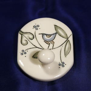 May include: White ceramic hook with a blue bird perched on a branch with green leaves and blue berries. The hook is shaped like a rounded rectangle.