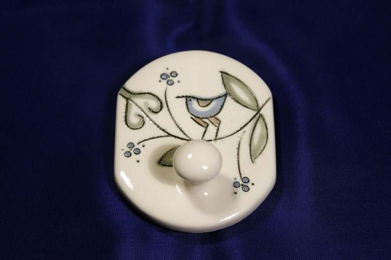 May include: White ceramic hook with a blue bird perched on a branch with green leaves and blue berries. The hook is shaped like a rounded rectangle.