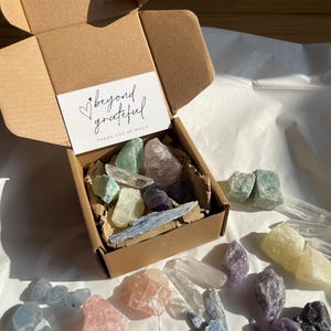 Pode incluir: Uma caixa de cartão aberta cheia de vários cristais coloridos e um pequeno cartão que diz "beyond grateful". Os cristais incluem quartzo transparente, quartzo rosa, ametista e cianita azul. Os cristais estão dispostos sobre uma superfície branca.
