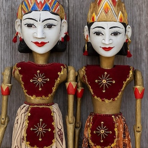 以下が含まれることがあります： 顔が描かれ、赤い唇と精巧な頭飾りを持つ2つのインドネシアの木製人形。 赤と金の装飾が施されたトップスと柄物のスカートを着用しています。 人形は木製の背景に展示されています。