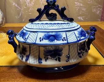 May include: A white ceramic serving dish with a blue and white floral pattern. The dish has a lid with a decorative handle and two handles on the sides.