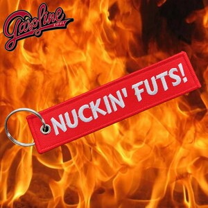 May include: Red keychain with white embroidered text that reads "NUCKIN' FUTS!" The keychain is against a background of flames.