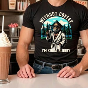 May include: Black t-shirt with a graphic of Bigfoot wearing sunglasses and a robe, holding a coffee cup. The text reads "WITHOUT COFFEE I'M KINDA BLURRY" in white. The design includes mountains and trees.