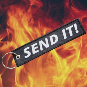 May include: Black rectangular keychain tag with the embroidered text "SEND IT!" in white. The tag has a silver metal ring attached. The background is a fiery orange and yellow.