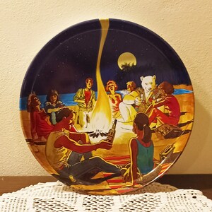 May include: A round, vintage metal tray with a painted scene of people gathered around a campfire on a beach at night. The tray has a dark blue background with a full moon and stars. A white crocheted doily is beneath the tray.