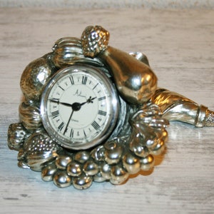 Può includere: Orologio argentato con quadrante rotondo e indici delle ore in numeri romani. L'orologio è circondato da un gruppo di frutta e verdura scolpite, tra cui uva, una pera e una cornucopia.
