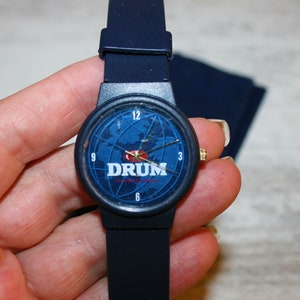 May include: A blue wristwatch with a black band and a blue face. The face has a world map design and the word "DRUM" printed on it.