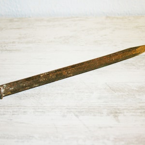 Puede incluir: Una herramienta de metal oxidada con un extremo puntiagudo y un mango. La herramienta está etiquetada con "HENSCHMIDT" y "L.F. SCHRIE" en el mango.