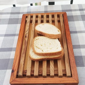 May include: A rectangular wooden bread board featuring a slatted design with a raised frame. Three slices of white bread with a golden-brown crust are stacked in the center of the board, resting on the parallel wooden slats of the tray.