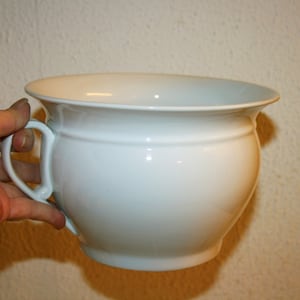 Peut inclure: Un pot de chambre en céramique blanche avec une seule poignée. Le pot est rond et a une finition lisse et brillante.