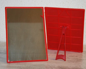 May include: Two red plastic mirrors with a stand. The mirror on the left is a standard mirror, while the mirror on the right has a grid pattern.
