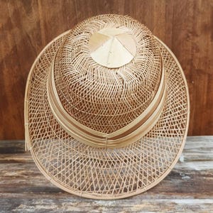May include: A light brown woven hat featuring a wide brim and a structured crown. The hat is constructed from thin, flexible strips of natural material arranged in a cross-hatched pattern, with a solid circular piece at the very top centre.
