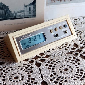 May include: A vintage Sharp digital alarm clock with a cream-colored case and a silver-toned face. The display shows the time as 2:27 PM, Monday. The clock has several buttons for setting the time and alarm.