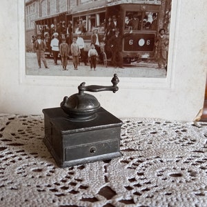 Può includere: Un macinacaffè vintage in metallo con una manovella e un cassetto. Il macinacaffè è dipinto di un colore grigio scuro e ha un aspetto usurato e antico.