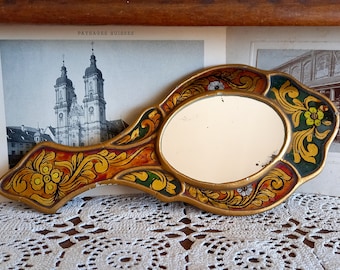 Puede incluir: Un espejo de mano de madera pintado a mano con un marco dorado y diseños florales en tonos rojos, amarillos y verdes. El espejo tiene una forma ovalada y está rodeado por un borde decorativo.