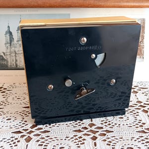 May include: A black music box with a gold trim. The music box has a small, round, silver knob on the front. The music box has a label on the front that reads 'root 8220-8311x'.